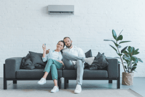 Male and Female Sitting closely and smiling on dark grey sofa with Ac unit mounted on wall behind them